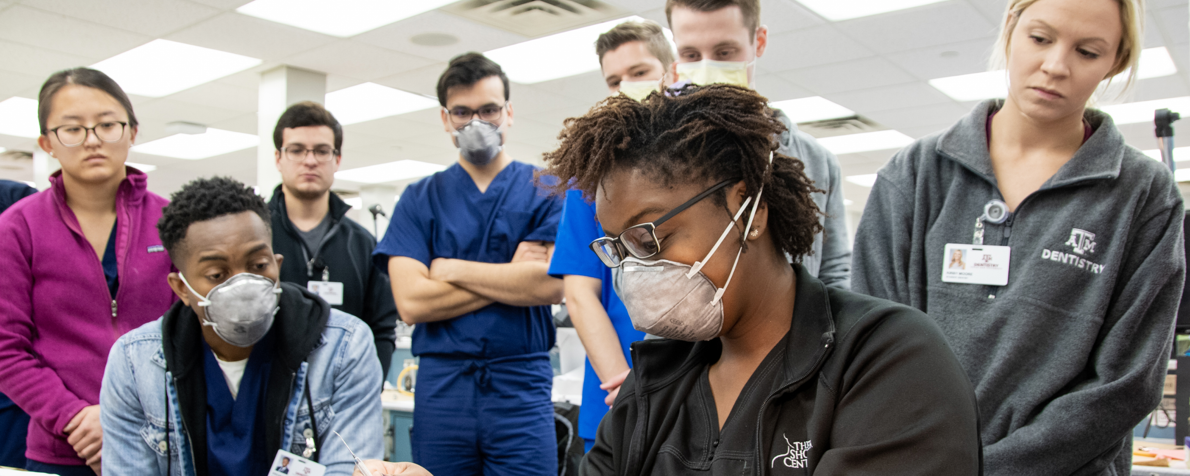 A group of dental students observe a demonstration in a clinical lab setting.