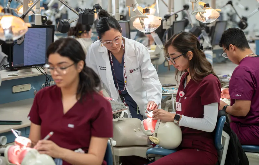 A dental hygiene instructor in a white coat supervises students practicing on manikins in a simulation lab