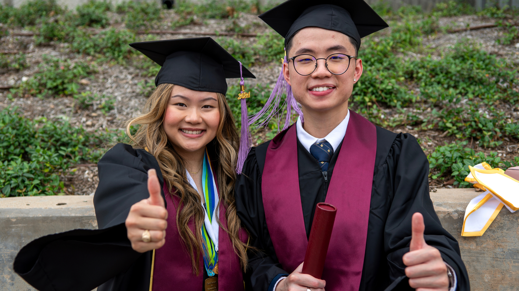 Two smiling dental school graduates in caps and gowns give thumbs up after commencement.