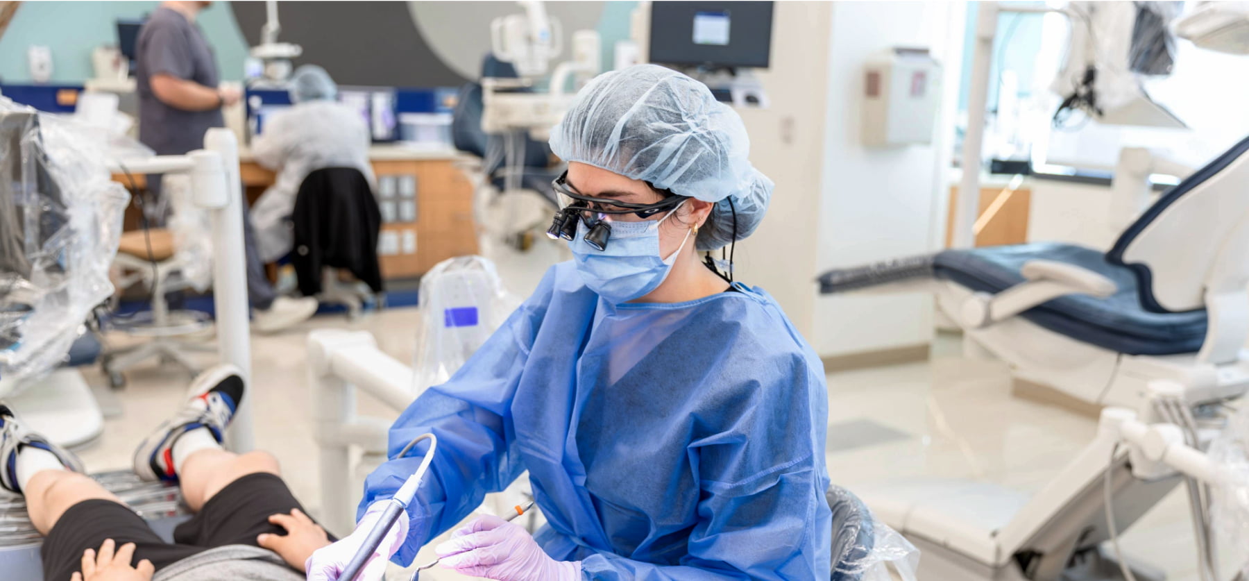A dental student in full clinical attire performs a procedure on a patient in a dental school clinic.