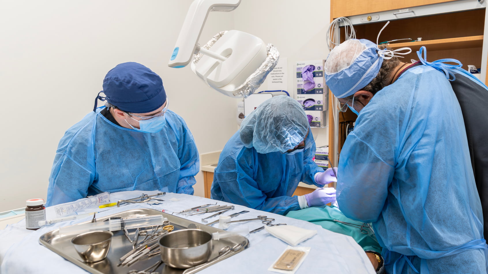 A surgical team in full protective gear performs an oral surgery procedure with a tray of instruments in the foreground.