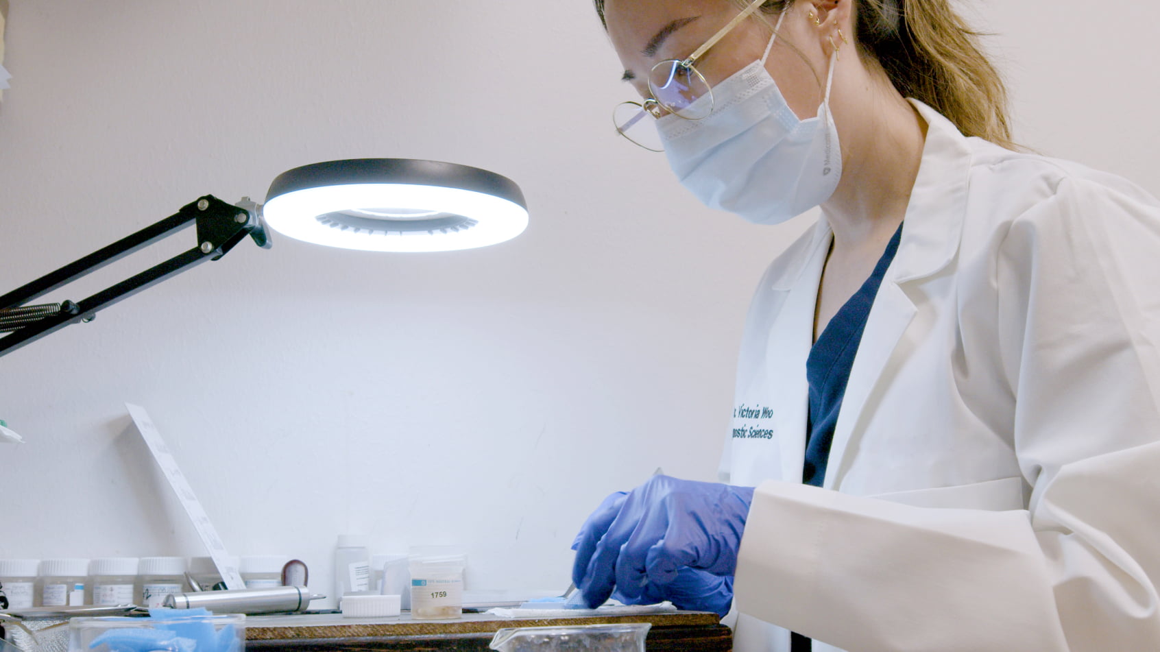 A pathology student or researcher in a white coat examines specimens under a magnifying lamp.