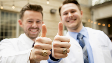 Two smiling dental students in white coats give thumbs up.