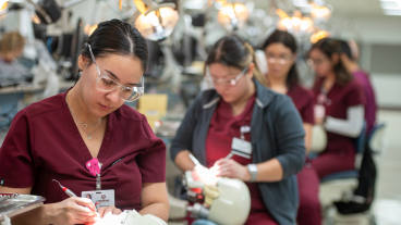 A row of dental students in scrubs practice clinical skills in a dental simulation lab.