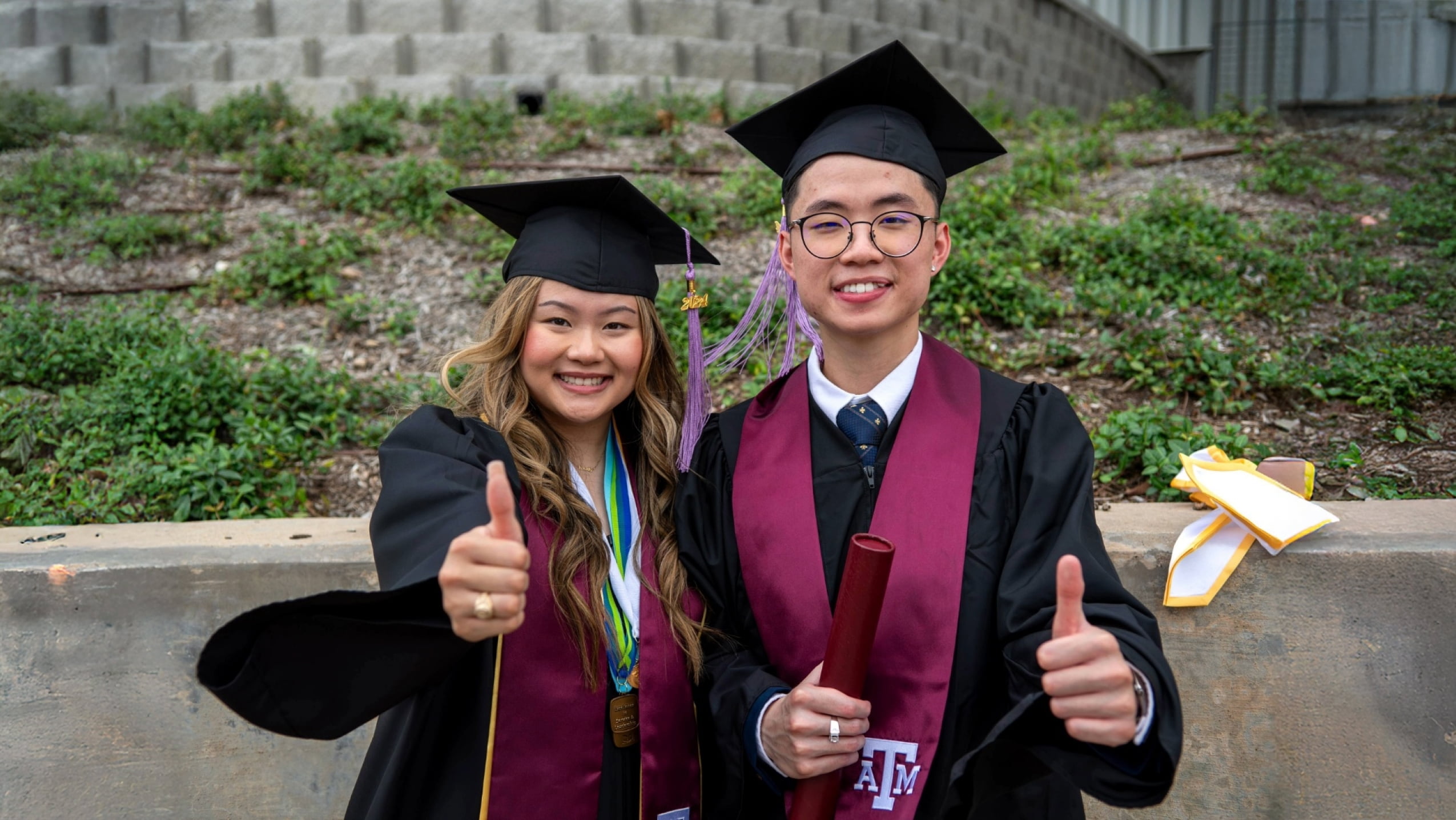 Two smiling dental school graduates in caps and gowns give thumbs up after commencement.