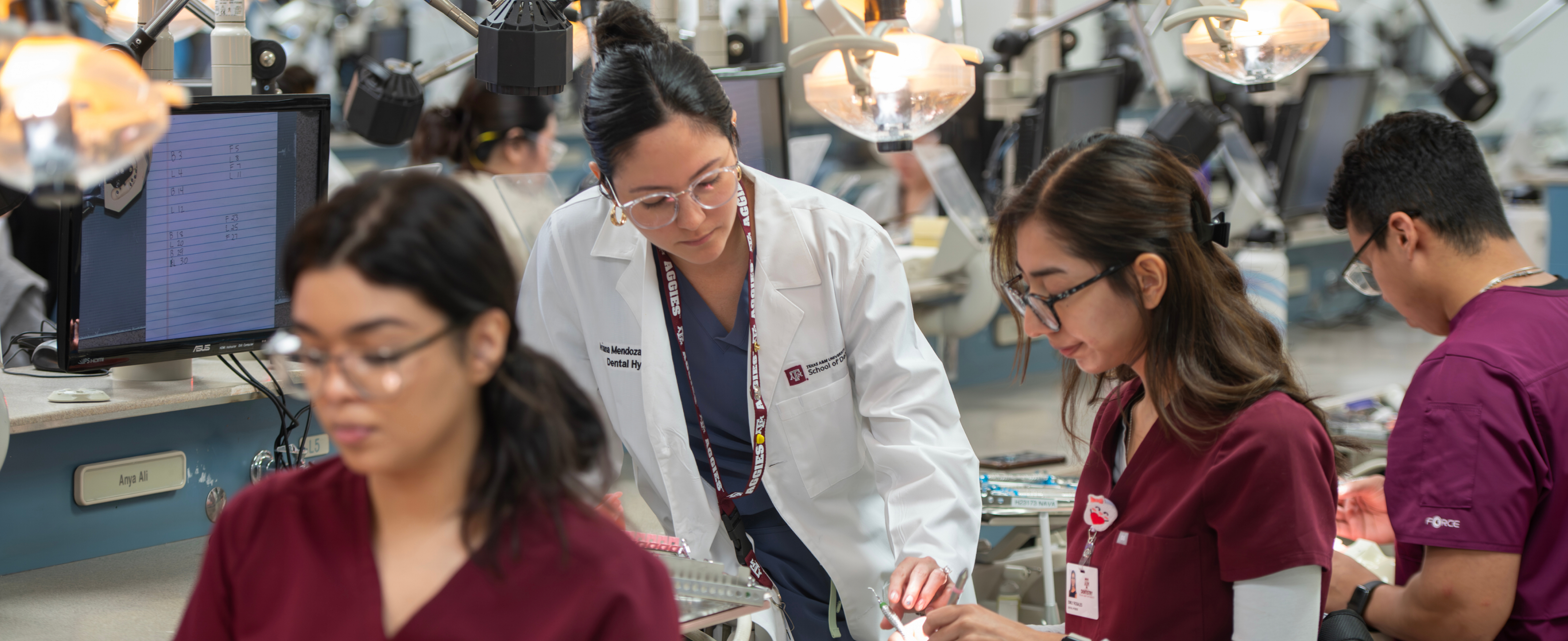 A dental hygiene instructor in a white lab coat guides a student in maroon scrubs during a practical session in a high-tech dental simulation lab.