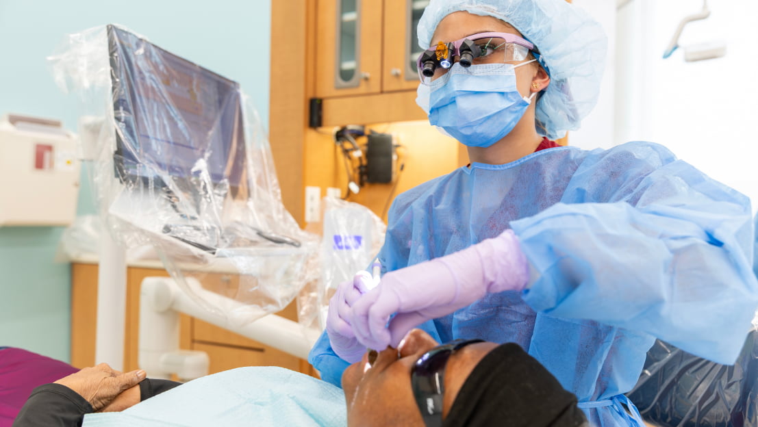 A dental hygiene student in a blue gown, hair net, mask, and loupes glasses performing a dental procedure on a patient in a clinic chair.