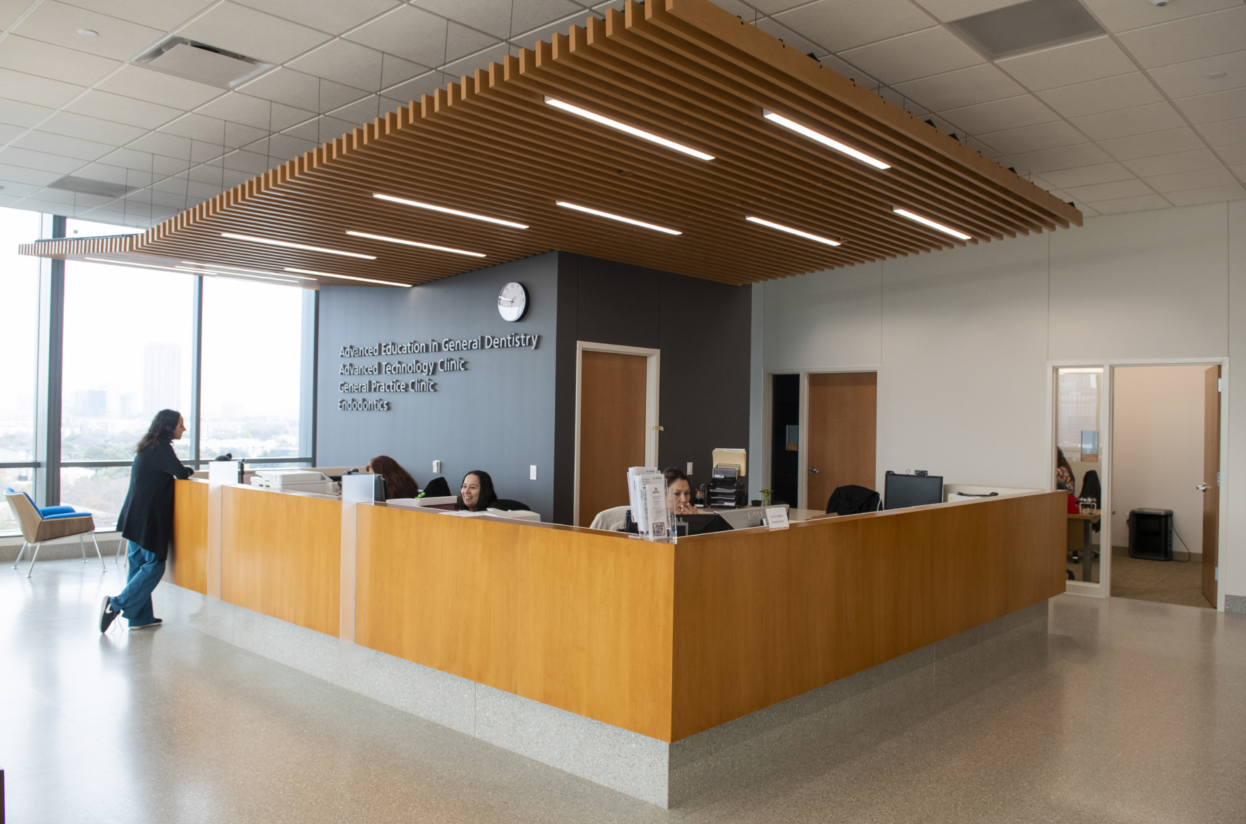 A modern reception desk at a dental clinic with staff assisting patients under a wood-slat ceiling with linear lighting.