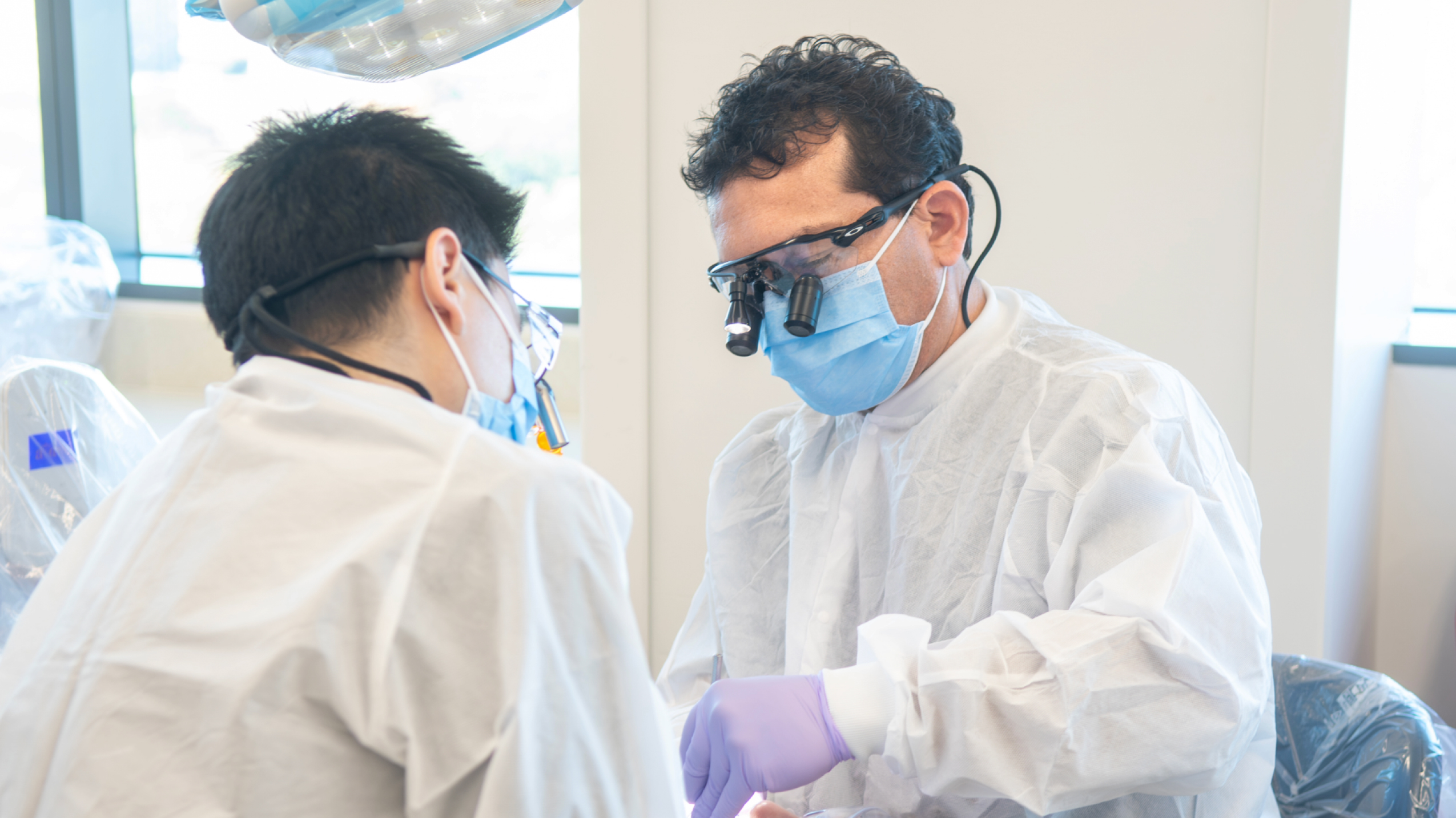 Dental practitioners in full protective gear and surgical loupes focused on a patient procedure in a clinical setting.