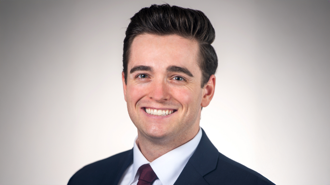 Profile headshot of Nathan Baker wearing a suit and maroon tie, smiling against a gray background.