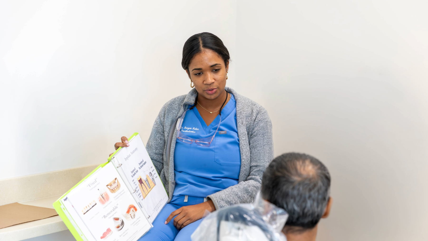 A prosthodontics resident showing an educational binder with dental implant diagrams to a patient during a consultation.
