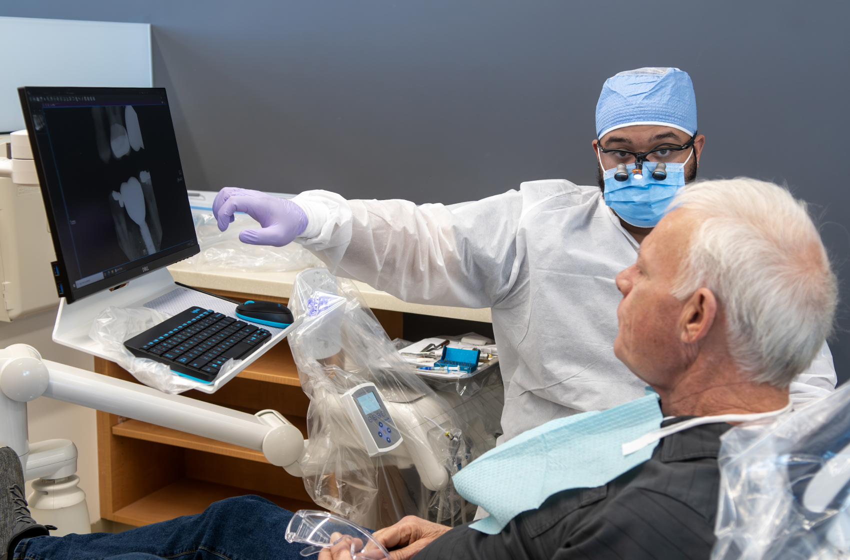 A dental provider reviews X-ray images on a monitor with an adult patient seated in a dental chair.