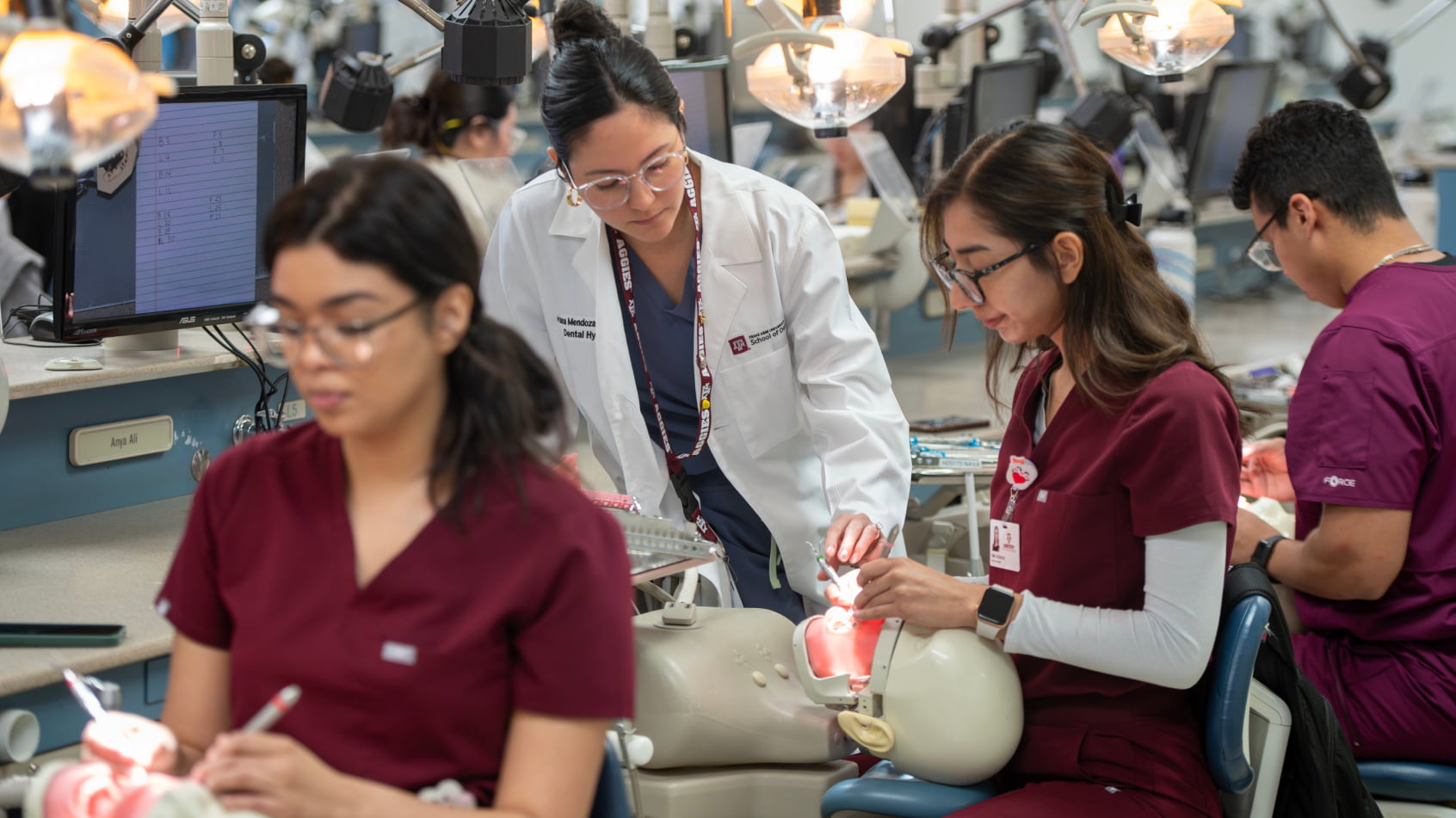 A faculty instructor in a white lab coat mentoring a dental student working on a patient simulation mannequin in a dental lab.