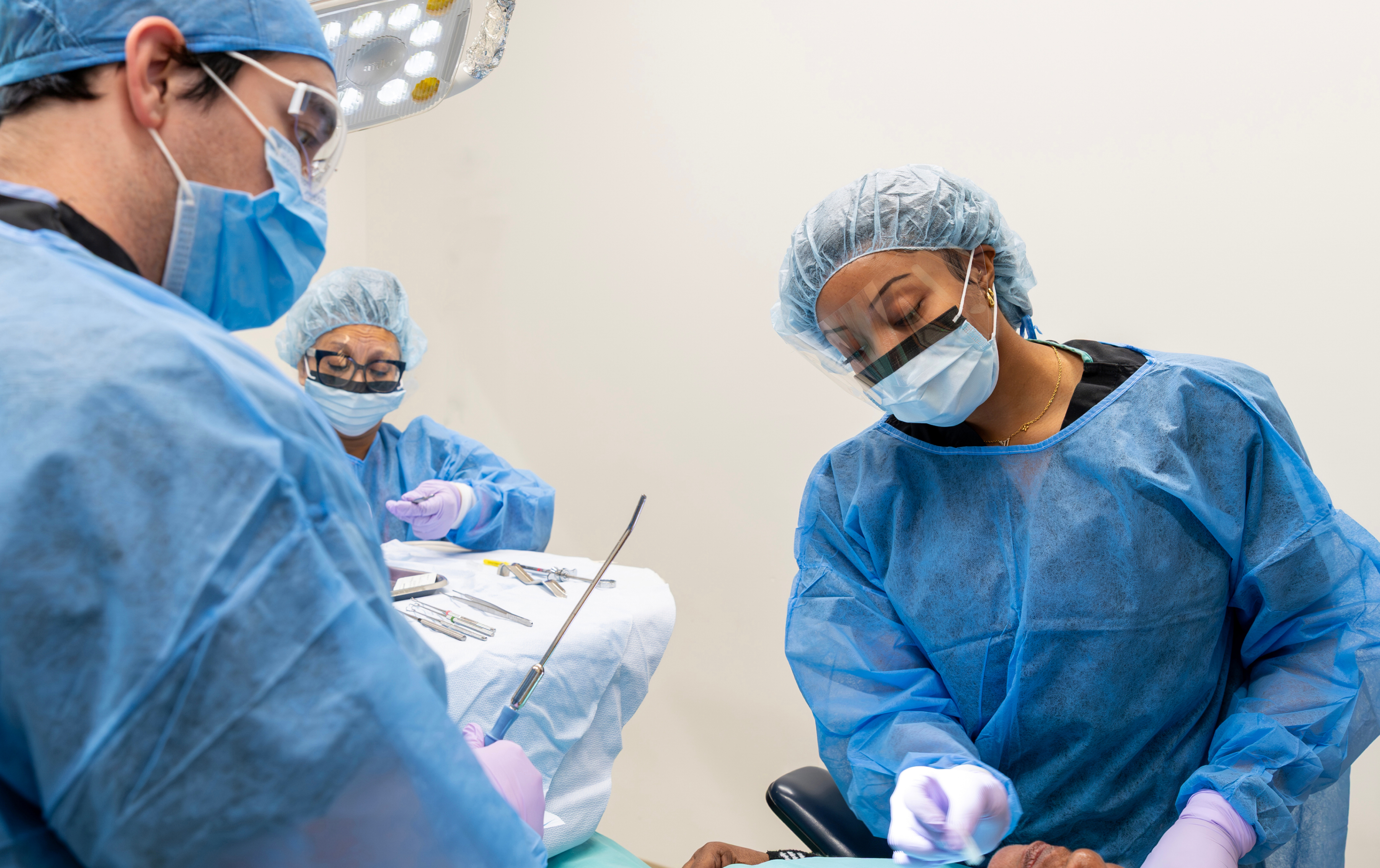 Three dental surgical practitioners wearing sterile gowns, masks, and face shields working in tandem during a clinical procedure.