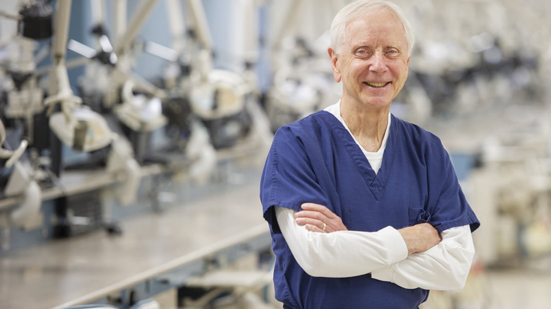 A smiling senior faculty member wearing blue scrubs with his arms crossed in a dental clinic.