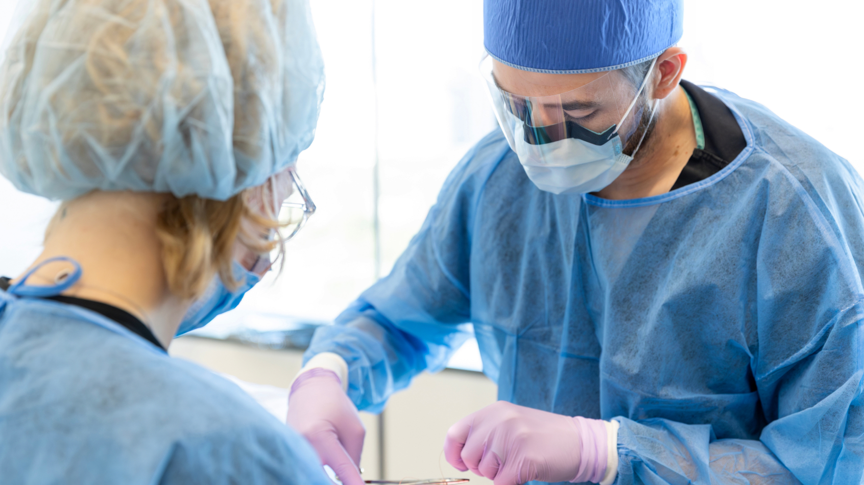 Two dental surgical practitioners in full sterile protective gear and face shields intently focused on a clinical procedure.