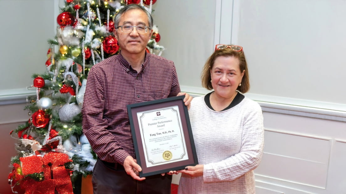 Feng Tao, M.D., Ph.D., smiling alongside a colleague while holding his framed Premier Performance Award certificate in front of a decorated holiday tree.