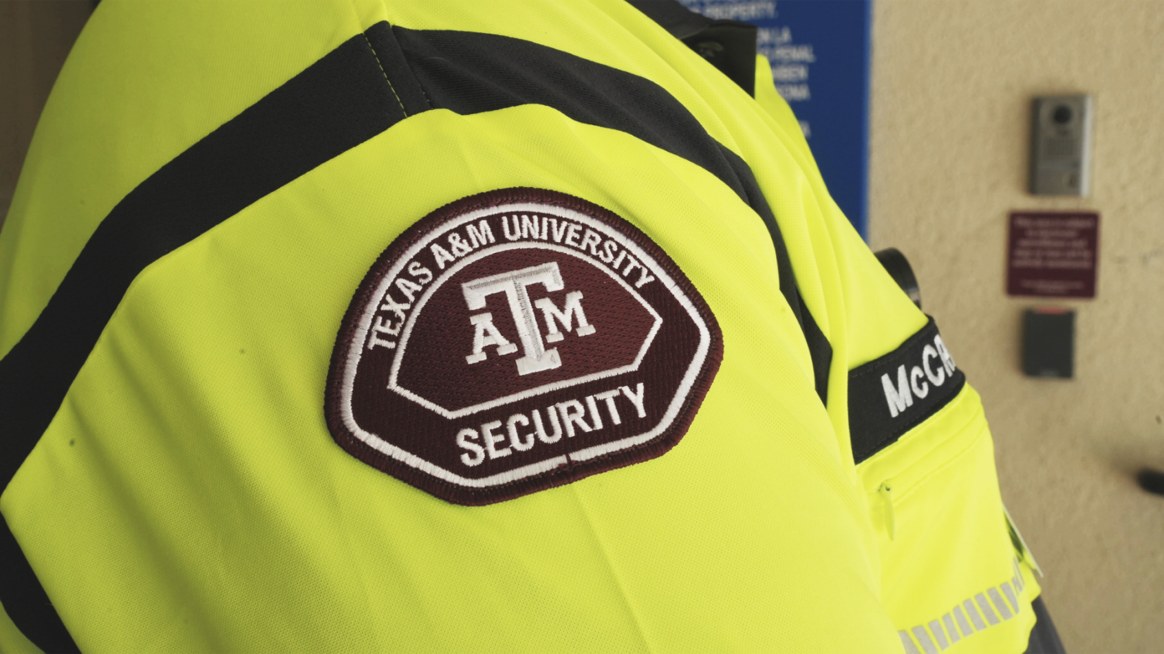 A close-up view of a Texas A&M University Security uniform, focusing on the official brown and white patch stitched onto a bright neon-yellow security vest.