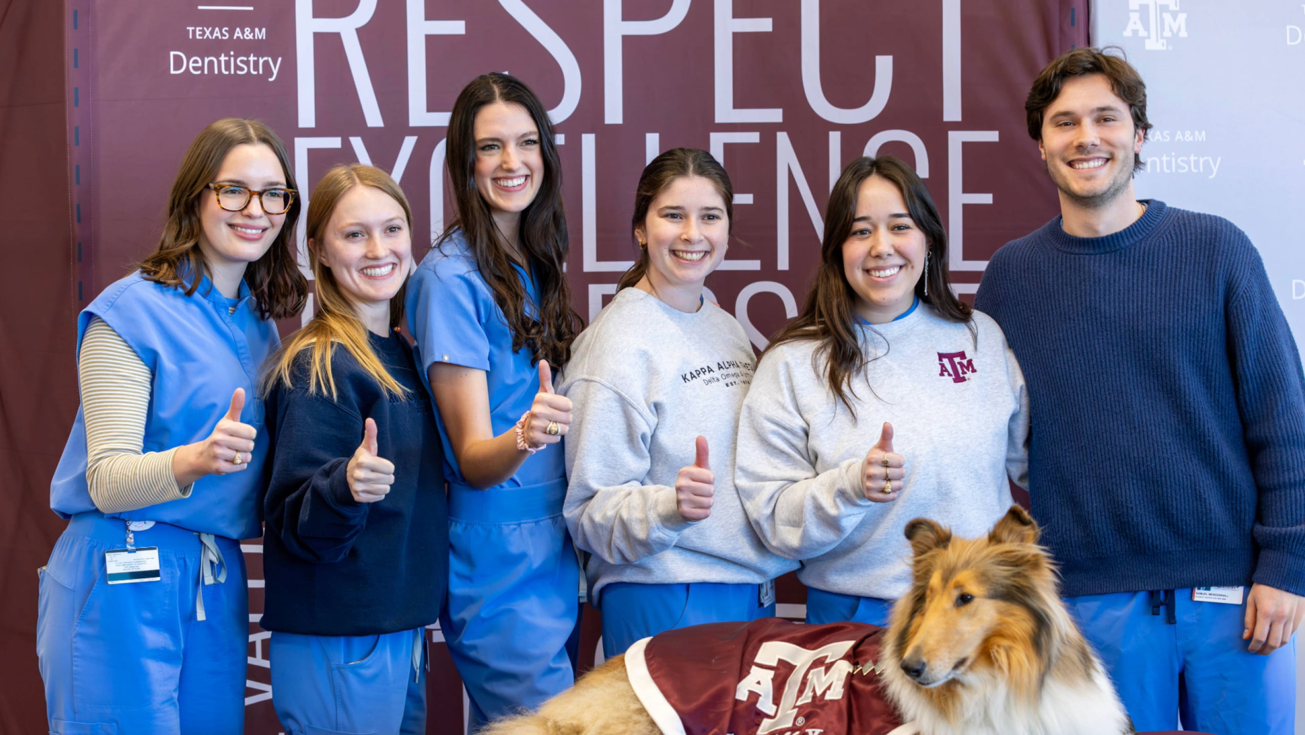 A group of five smiling dental students in blue scrubs and gray Texas A&M sweatshirts giving a thumbs-up while posing indoors with Reveille, the Rough Collie mascot.