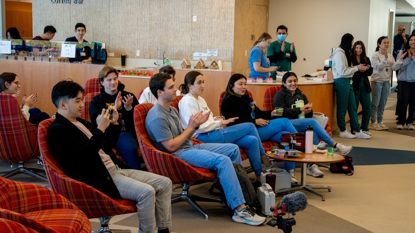 A group of dental students sitting in red lounge chairs in a campus common area, smiling and clapping during a presentation or event.