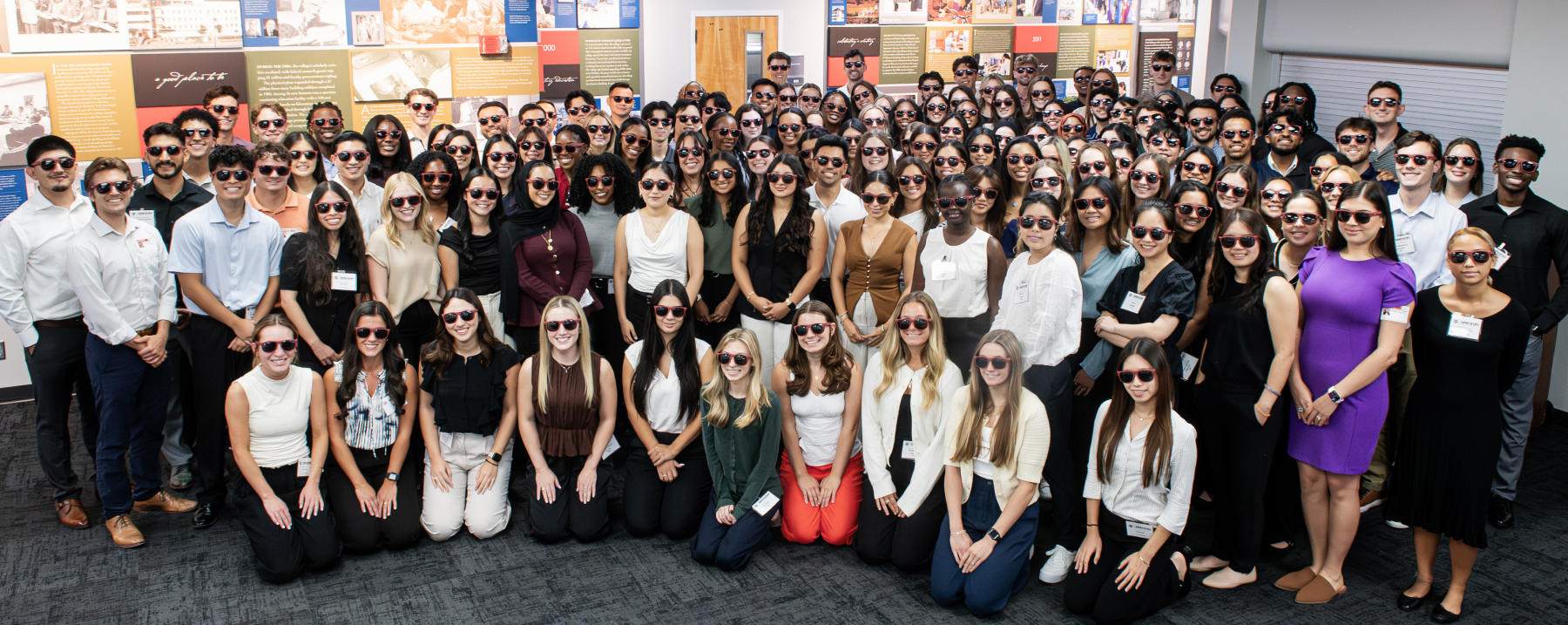 A large group photo of dozens of dental students, faculty, or staff members all wearing sunglasses and smiling together indoors in front of a campus display wall.