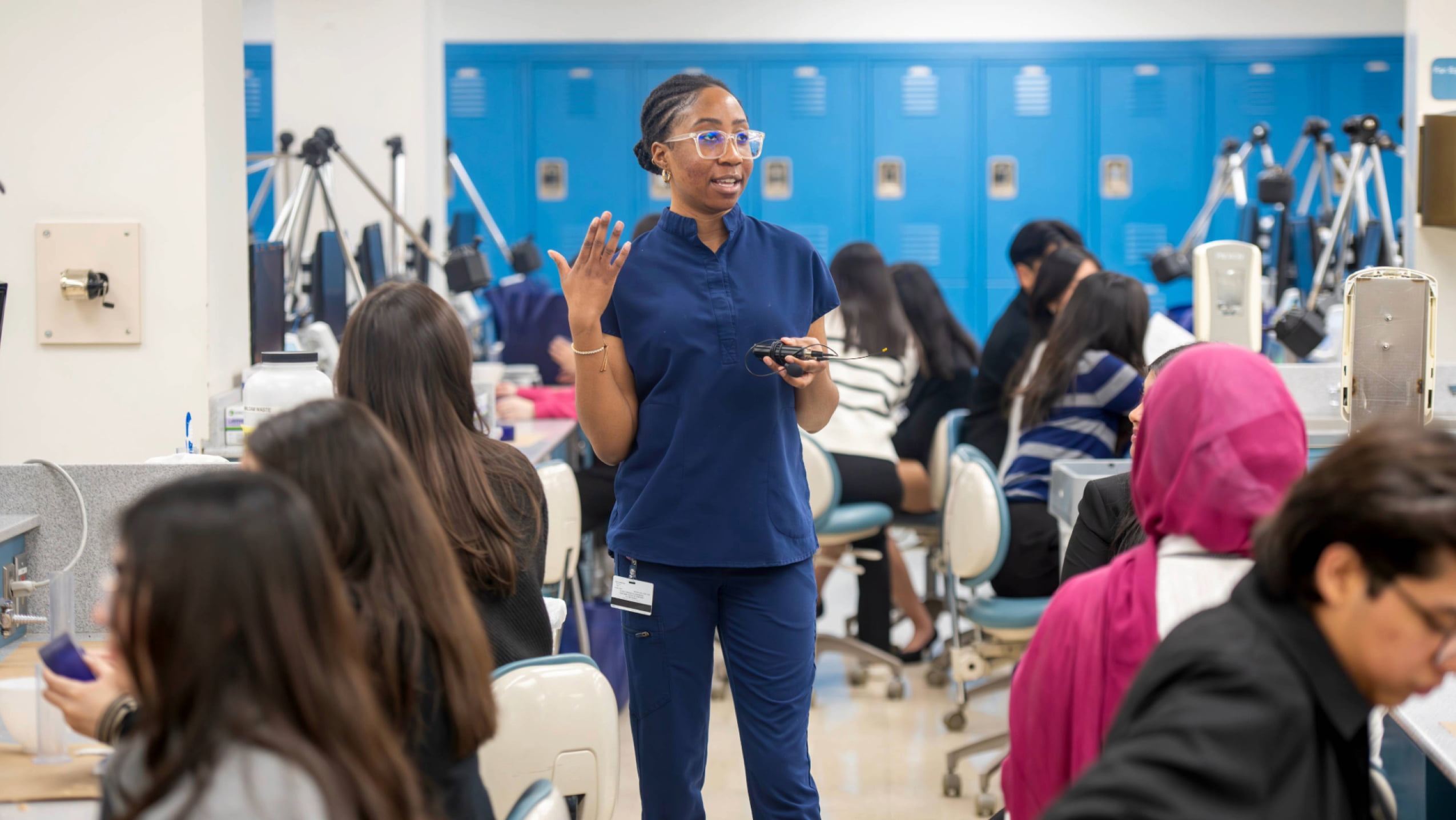 A dental student in blue scrubs gestures while speaking to classmates in a dental simulation lab with blue lockers in the background.