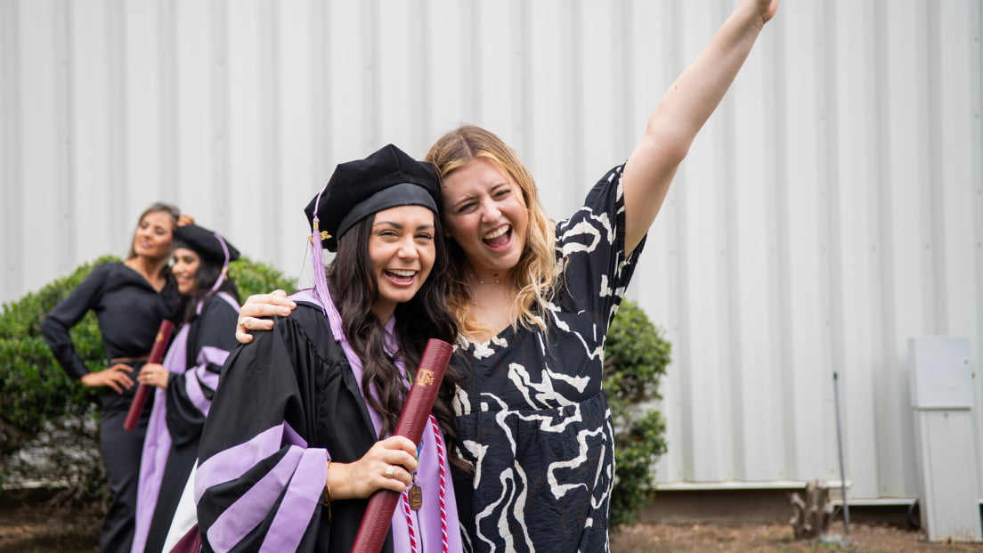A happy dental school graduate holding her diploma scroll poses and celebrates with an excited friend outdoors after a commencement ceremony.