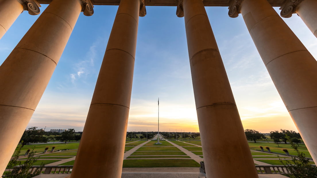 Looking out through the large stone columns of the Texas A&M Administration Building onto the campus quad at sunset.