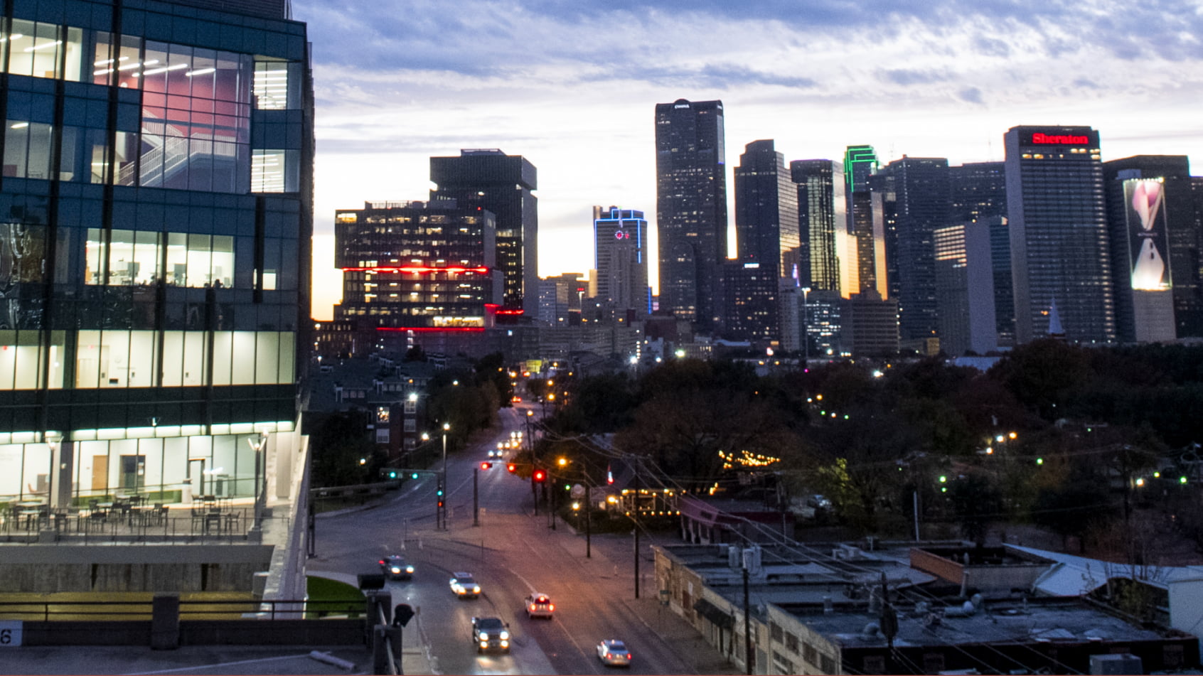 A dusk view of the downtown Dallas skyline and city streets seen from the modern, lit glass building of the Texas A&M School of Dentistry.