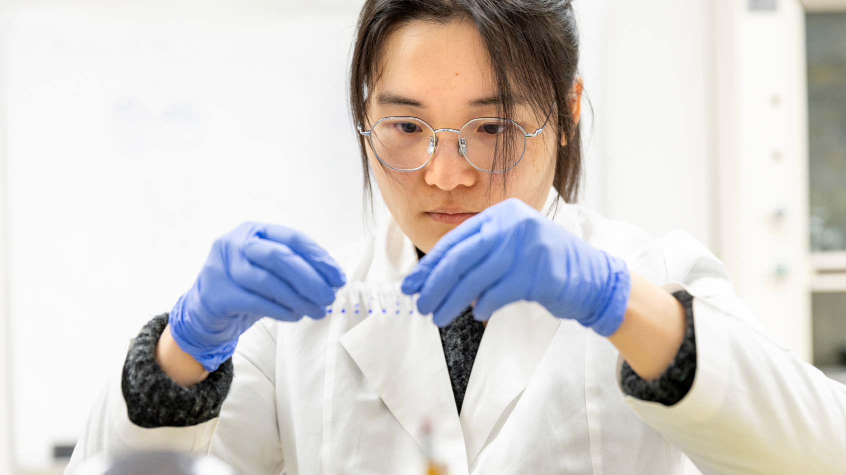 A close-up view of a female researcher in a white lab coat, glasses, and blue gloves carefully examining a strip of microcentrifuge tubes in a laboratory setting.