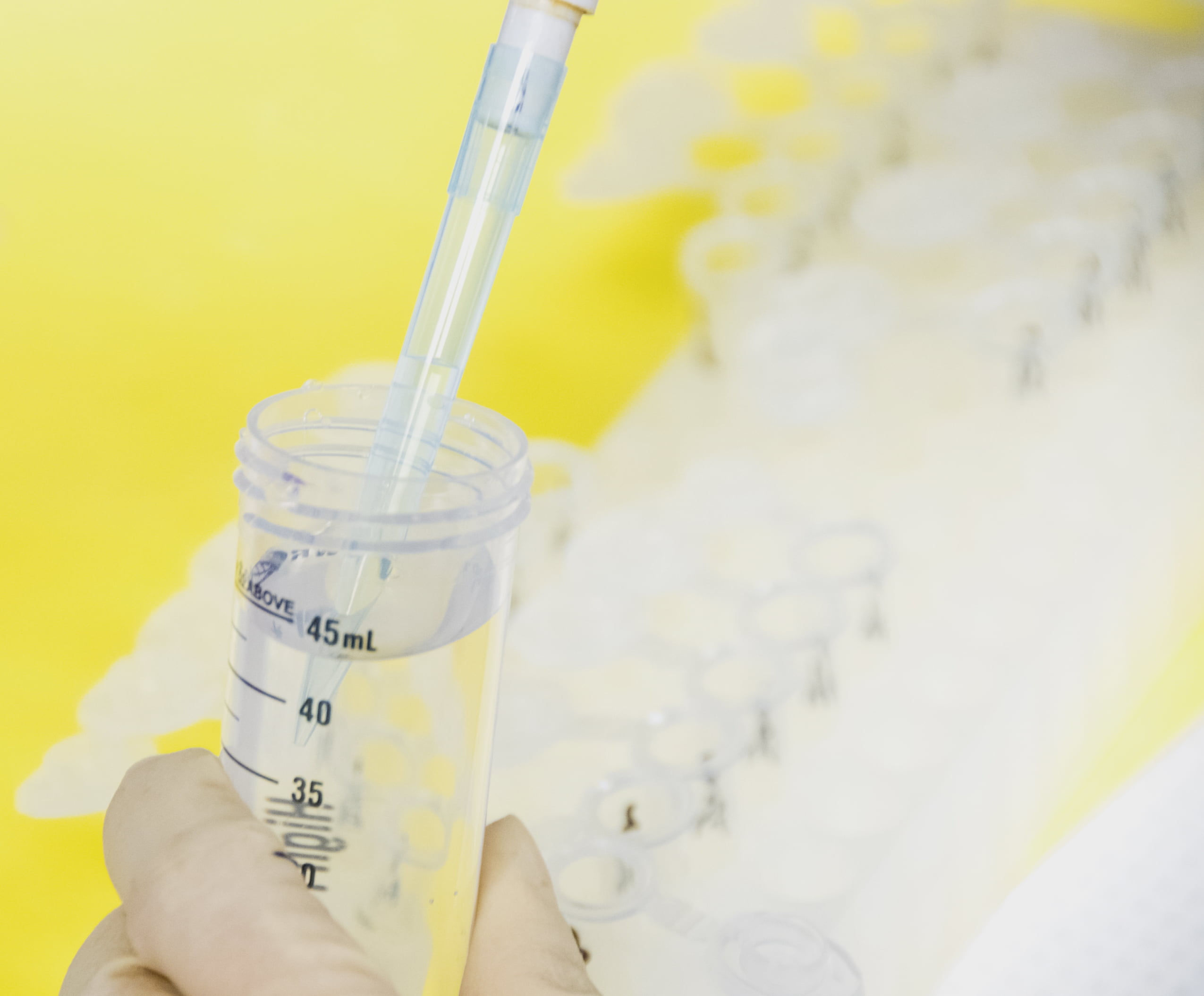 Close-up of a clear plastic laboratory pipette transferring liquid into a graduated 45 milliliter centrifuge tube against a vibrant yellow background.