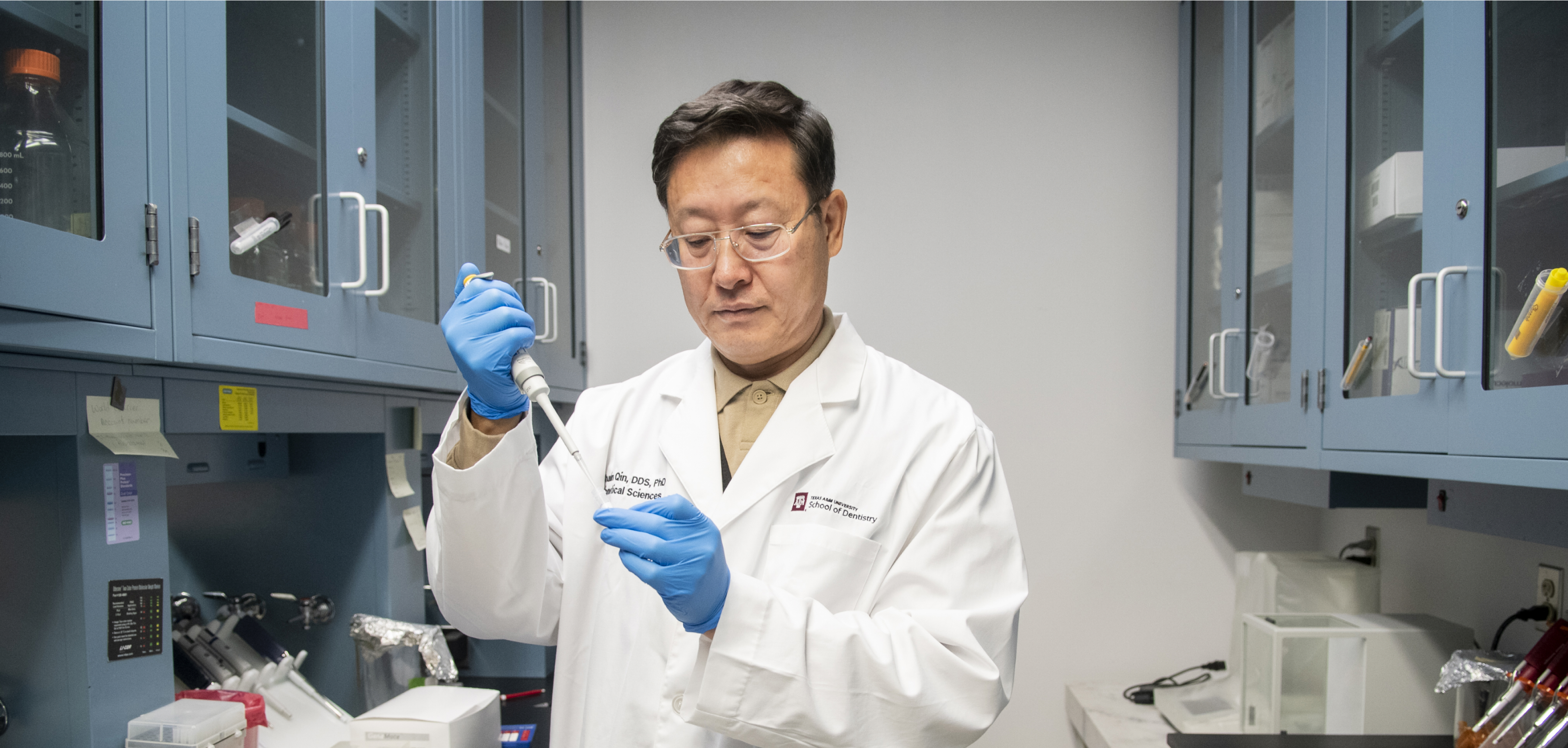 A male researcher in a white lab coat and blue gloves using a pipette in a laboratory setting.